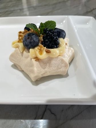 Mini pavlova meringue dessert topped with lemon cream, fresh blueberries and blackberries, a mint leaf and crunchy nut crumble on a white square plate