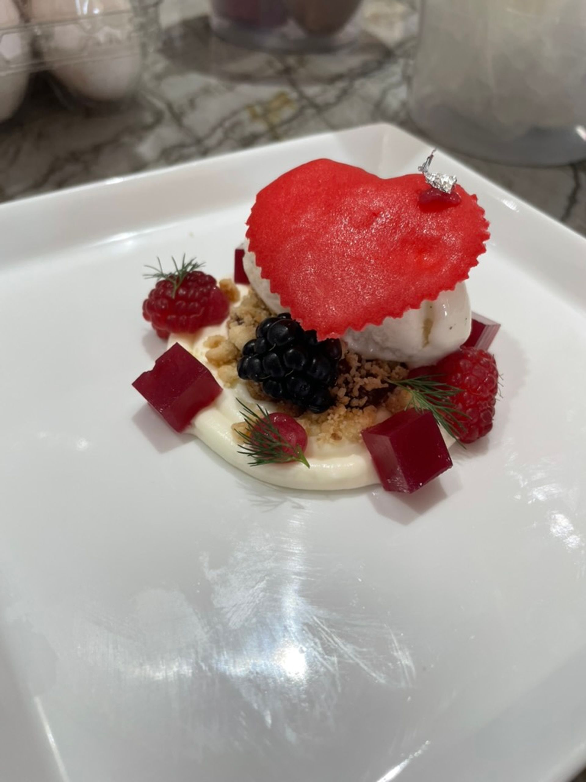 Fine-dining plated dessert on a white square plate: vanilla ice cream topped with a red heart-shaped tuile, blackberry and raspberries, red gelatin cubes, creamy sauce and crumble garnish.