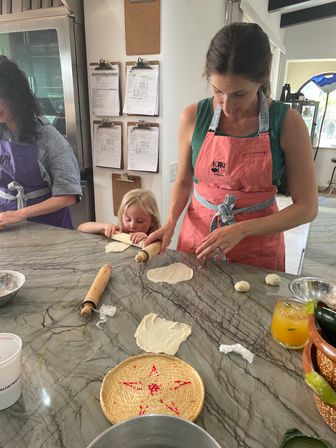 Woman and child rolling dough on a marble countertop during a hands-on cooking class, wearing aprons with rolling pins, dough rounds and kitchen tools visible.