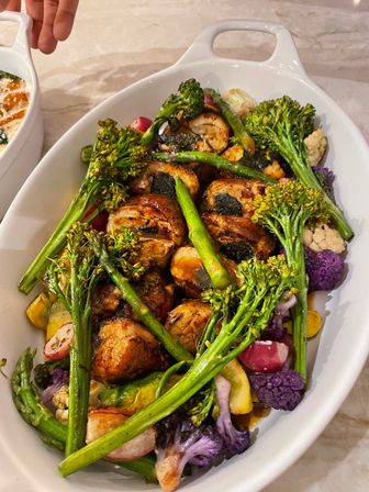 Oval white serving dish of seared scallops glazed in sauce with roasted seasonal vegetables — broccolini, asparagus, purple cauliflower and radishes.