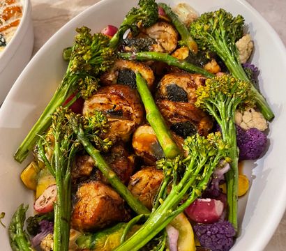 Oval white serving dish of seared scallops glazed in sauce with roasted seasonal vegetables — broccolini, asparagus, purple cauliflower and radishes.