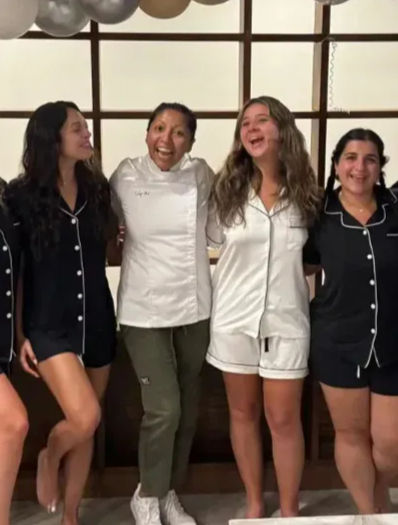 Four women smiling arm-in-arm at an indoor pajama party under metallic balloons — two in black pajama sets, one in white pajamas, and one in a white jacket and pants.