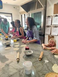 Bright community cooking class in a sunny kitchen — adults and a child in colorful aprons rolling dough on a marble countertop with rolling pins, bowls and drinks.
