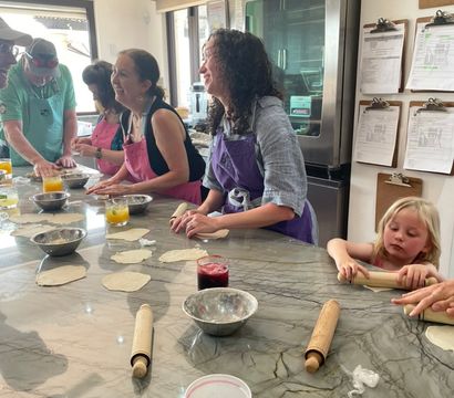 Bright community cooking class in a sunny kitchen — adults and a child in colorful aprons rolling dough on a marble countertop with rolling pins, bowls and drinks.