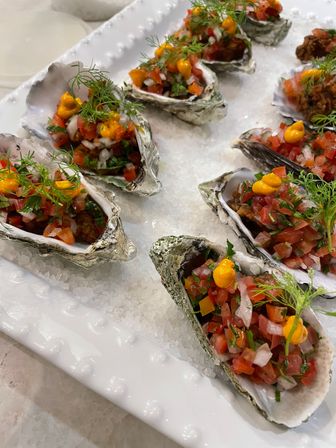 Bright oyster appetizers on a bed of coarse salt: half shells filled with diced tomato-and-onion salsa, orange aioli dots and fresh dill on a white platter.