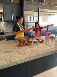 Two people in aprons preparing food at a marble kitchen island with a blender, bowls of fresh ingredients and stainless-steel ovens and refrigerator in the background.