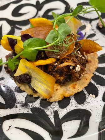 Mini tostada on a toasted corn tortilla topped with shredded braised meat, black beans, sesame seeds, crispy vegetable chips, edible flowers and pea-shoot microgreens on a black-and-white patterned plate