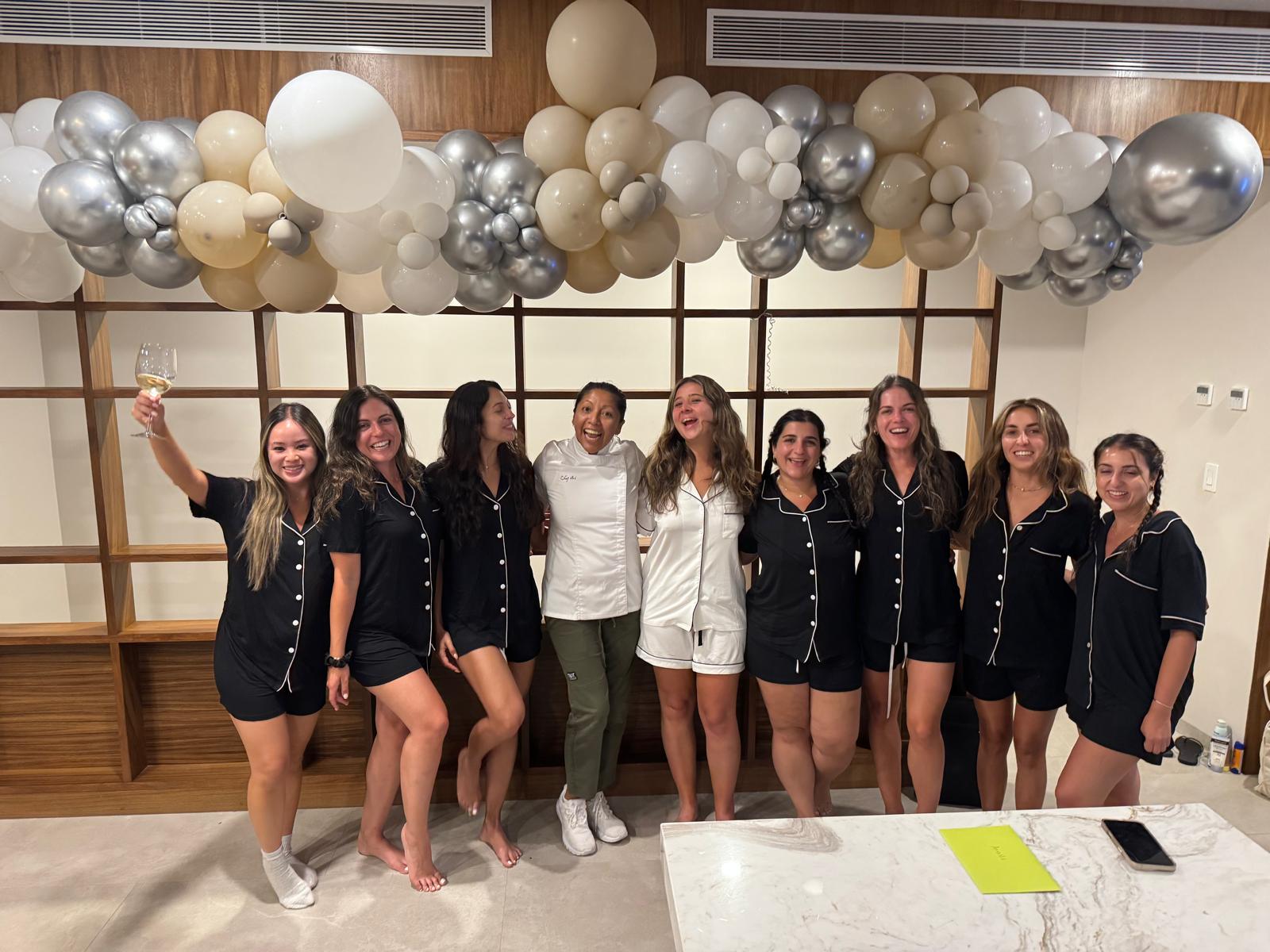 Group of friends in matching black and white pajamas celebrating indoors under beige and silver balloon garland, posing by a modern wood shelf and marble countertop while one holds a wine glass.