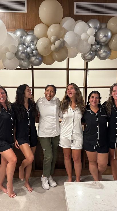 Group of friends in matching black and white pajamas celebrating indoors under beige and silver balloon garland, posing by a modern wood shelf and marble countertop while one holds a wine glass.