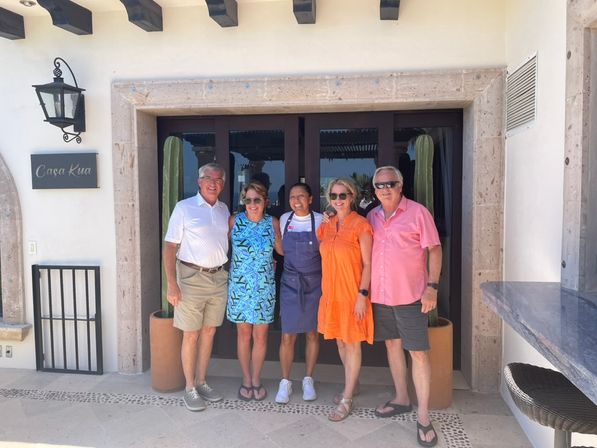 Five smiling adults in colorful summer outfits pose at the sunlit entrance of a white-stucco villa with stone trim and tall potted cacti — relaxed coastal vacation vibe.