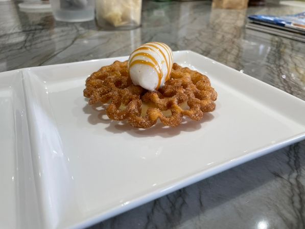 Crispy mini funnel cake dusted with cinnamon sugar topped with an oval scoop of vanilla ice cream and caramel drizzle, served on a white plate on a marble countertop.
