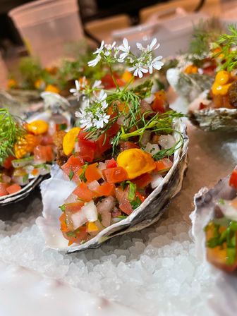 Raw oysters on the half shell topped with diced tomato-onion salsa, bright orange aioli dots, fresh herbs and white edible flowers, served on crushed ice — vibrant coastal seafood appetizer.