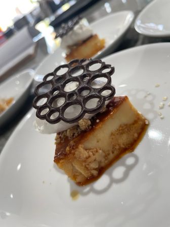 Caramel flan slice topped with whipped cream, crunchy crumble and a decorative chocolate lattice on a white plate — plated gourmet dessert.