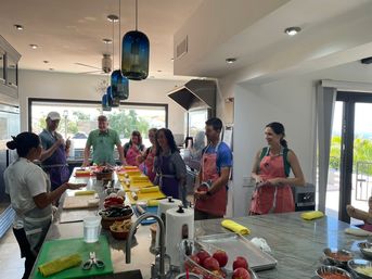 Sunlit ocean-view kitchen cooking class with adults in colorful aprons gathered around a long island with yellow cutting mats, rolling pins, bowls of fresh produce and blue pendant lights over a modern island.