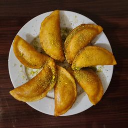 Golden fried dessert turnovers arranged in a circle on a white plate, dusted with crushed pistachios — crispy fried pastry on a dark wooden table