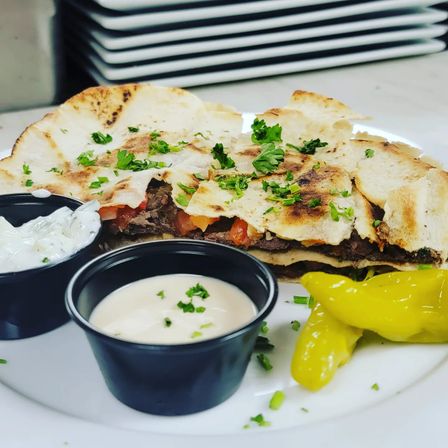 Grilled steak quesadilla on a white plate — charred flatbread filled with steak and tomatoes, garnished with chopped parsley, served with creamy dipping sauces and bright pepperoncini peppers.