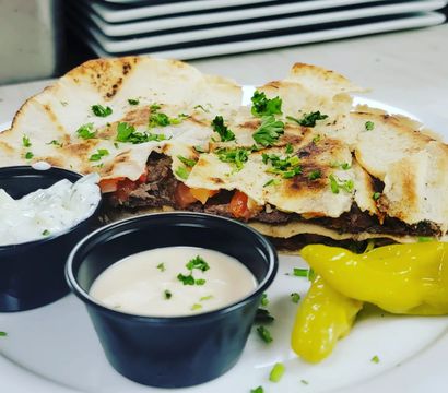 Grilled steak quesadilla on a white plate — charred flatbread filled with steak and tomatoes, garnished with chopped parsley, served with creamy dipping sauces and bright pepperoncini peppers.