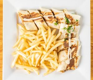 Golden French fries piled beside a sliced grilled tortilla wrap drizzled with creamy sauce and sprinkled parsley on a square white plate over a wooden table, casual plated lunch.