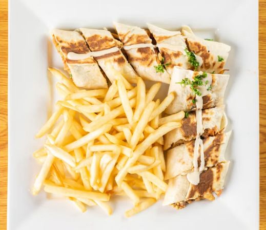 Golden French fries piled beside a sliced grilled tortilla wrap drizzled with creamy sauce and sprinkled parsley on a square white plate over a wooden table, casual plated lunch.
