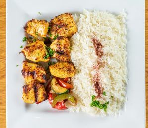 Grilled chicken skewers with charred bell peppers and zucchini alongside a mound of steamed long-grain rice sprinkled with spices and parsley on a square white plate on a wooden table.