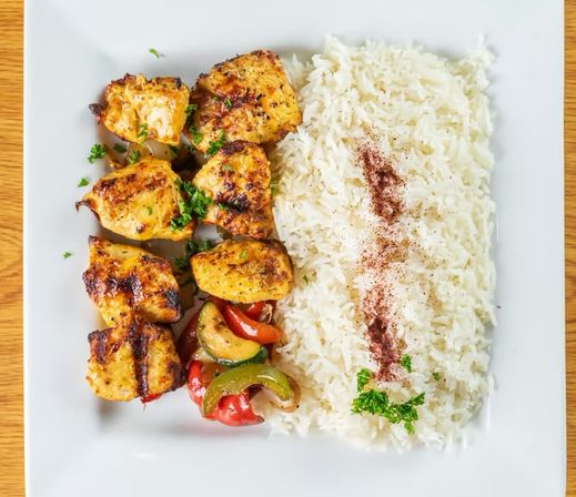 Grilled chicken skewers with charred bell peppers and zucchini alongside a mound of steamed long-grain rice sprinkled with spices and parsley on a square white plate on a wooden table.