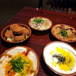 Mediterranean mezze spread on a dark wood table: five small clay bowls with falafel, creamy hummus drizzled with olive oil and paprika, yogurt labneh with za'atar, a bean salad, and an eggplant dip garnished with parsley — colorful Middle Eastern appetizers.