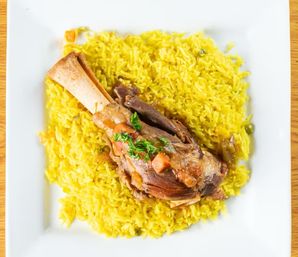 Overhead view of a braised lamb shank on saffron yellow rice, topped with chopped parsley and tomato on a white square plate.