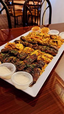 Mixed grilled kebab platter with marinated beef and chicken atop seasoned yellow rice, served with dipping sauce cups on a white platter on a wooden restaurant table.