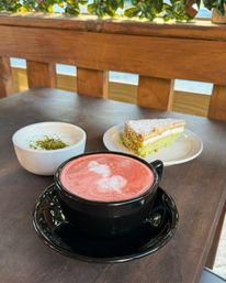 Pink frothy latte in a black cup on an outdoor café table, beside a small bowl of creamy dessert topped with chopped pistachios and a powdered sugar‑dusted green layered cake slice.