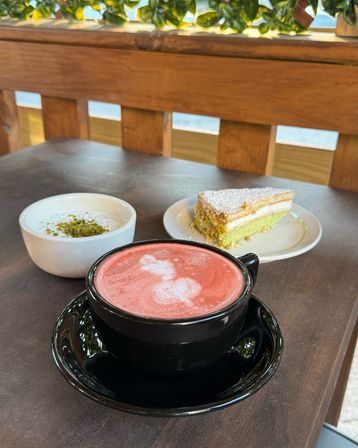 Pink frothy latte in a black cup on an outdoor café table, beside a small bowl of creamy dessert topped with chopped pistachios and a powdered sugar‑dusted green layered cake slice.