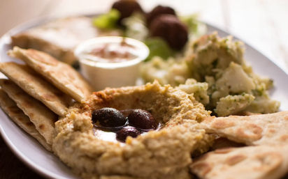 Mediterranean mezze plate with creamy hummus topped with black olives and olive oil, warm pita wedges, potato salad and falafel on a white plate