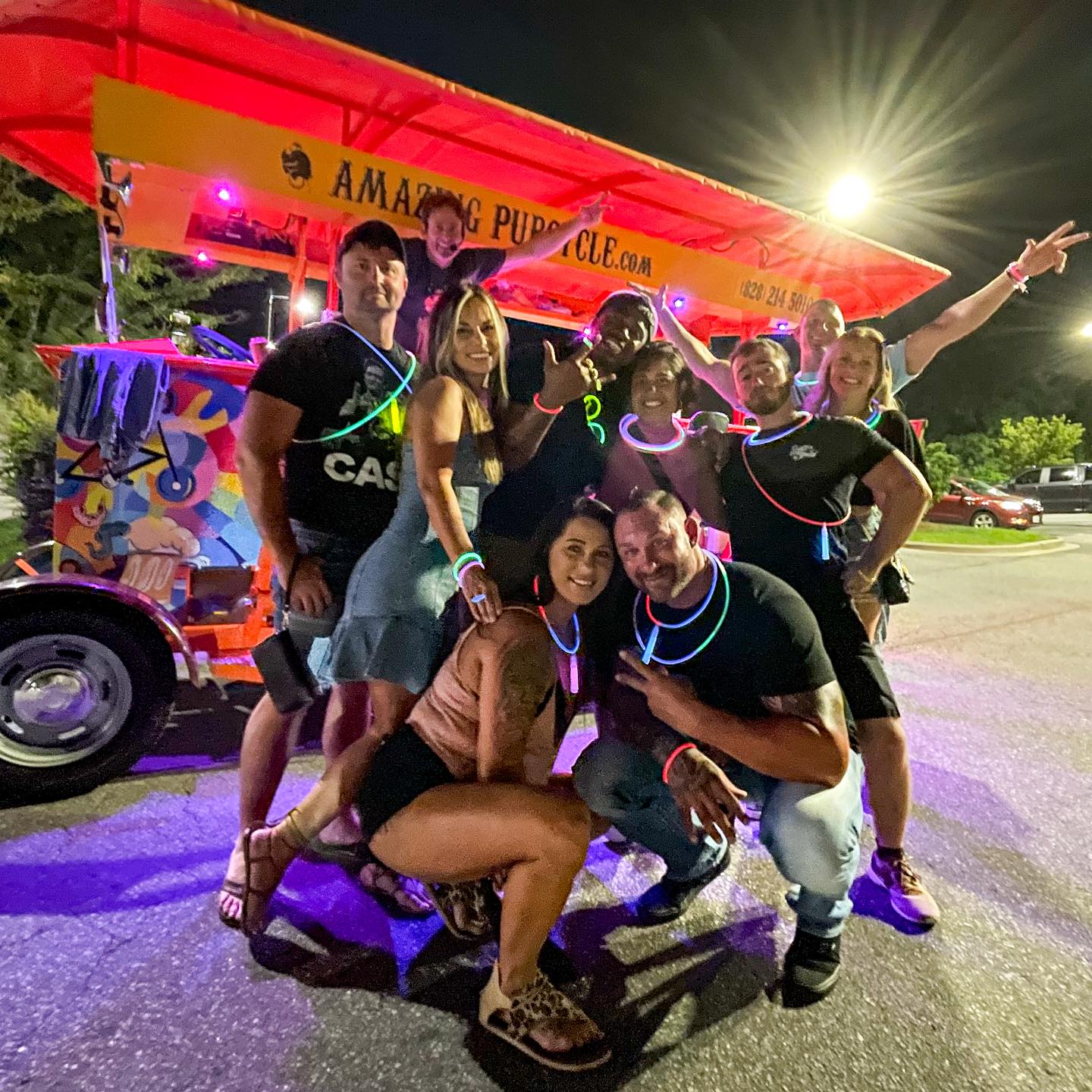 Lively group of friends at night posing around a colorful pedal party bike in a parking lot, wearing glow-stick necklaces and bracelets under a red canopy, smiling and flashing peace signs.
