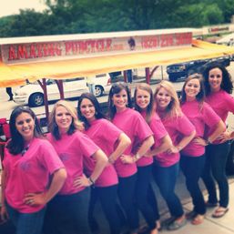 Eight smiling women in matching bright pink T-shirts and jeans stand in a row with hands on hips, posing in front of a yellow-canopied pedal party bike on a tree-lined city street.