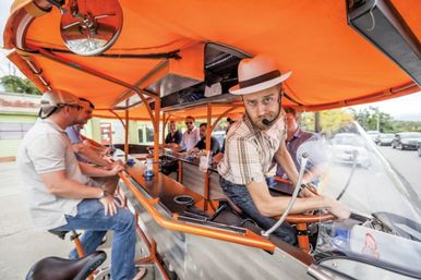 Pedal pub party bike with orange canopy: hat-wearing guide with headset steering while a group of riders sit at a bar-style counter on a sunny city street
