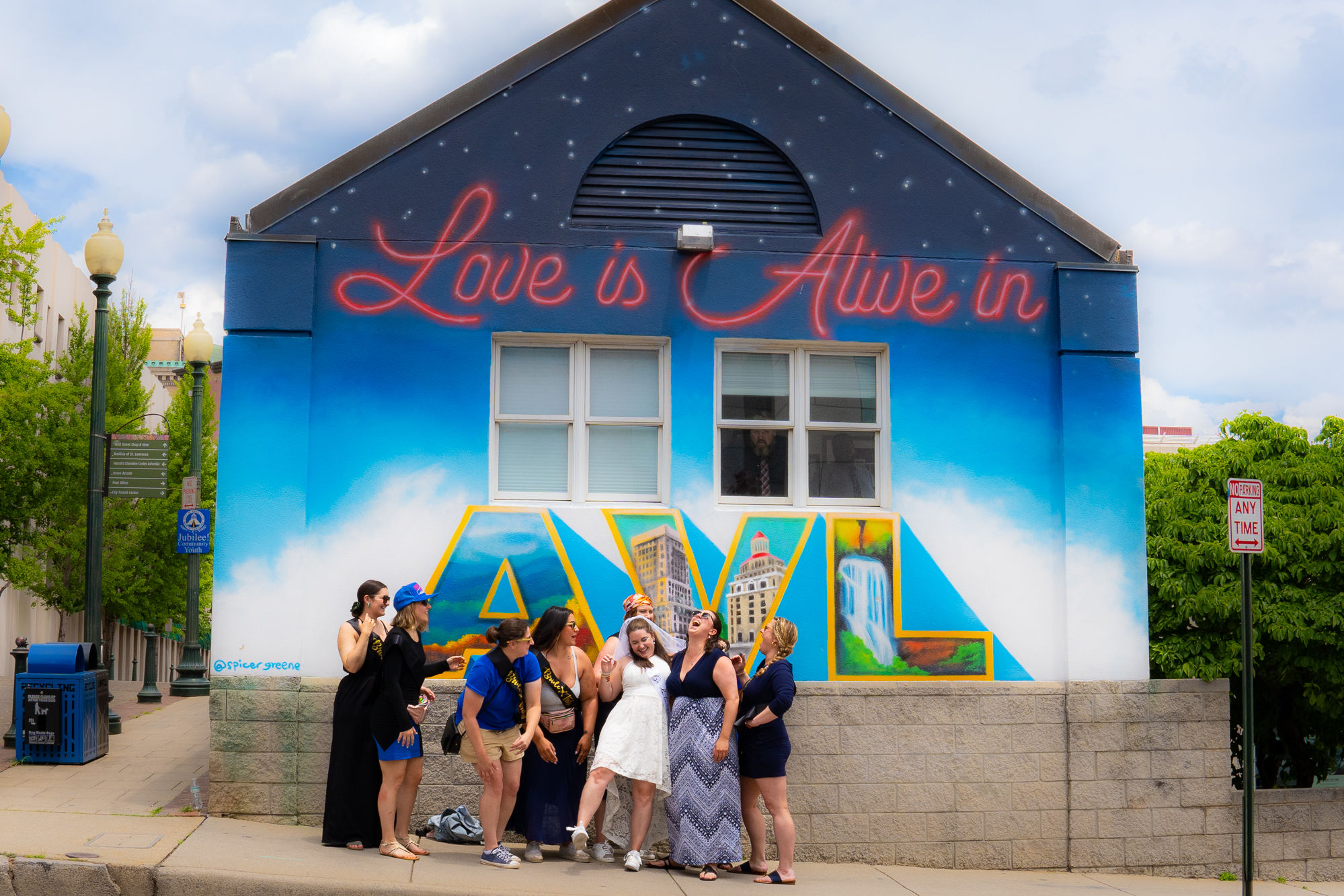 Group of women laughing and posing in front of an Asheville mural that reads "Love is Alive in AVL," bright blue sky background with large colorful cityscape letters on a building wall.