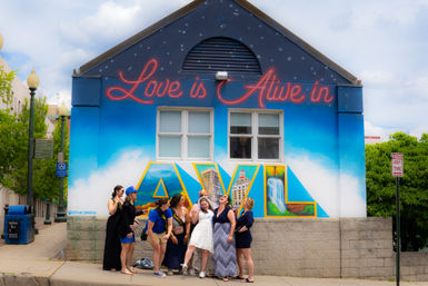 Group of women laughing and posing in front of an Asheville mural that reads "Love is Alive in AVL," bright blue sky background with large colorful cityscape letters on a building wall.
