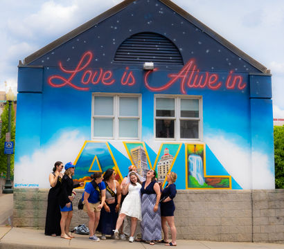 Group of women laughing and posing in front of an Asheville mural that reads "Love is Alive in AVL," bright blue sky background with large colorful cityscape letters on a building wall.