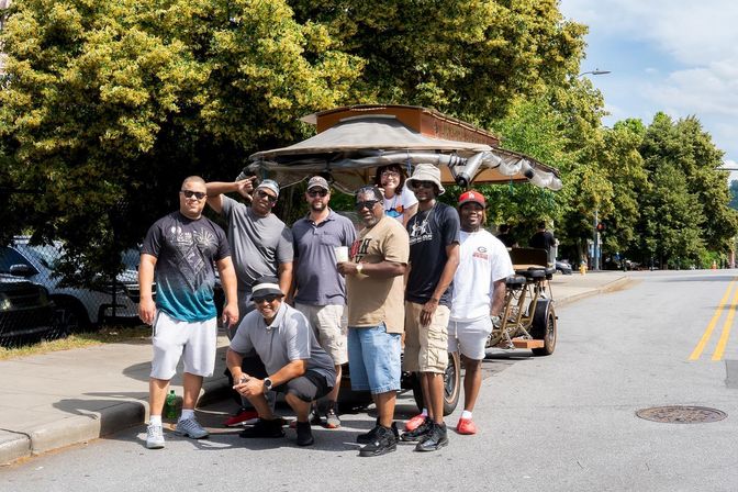 Eight men in casual summer clothes smiling and posing beside a pedal-powered party bike on a sunlit, tree-lined urban street.