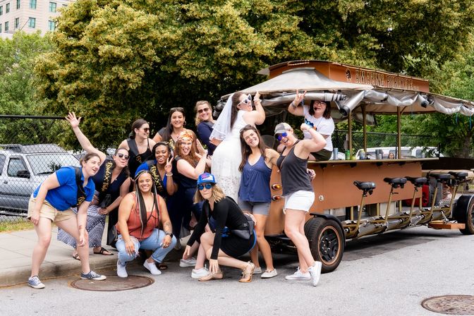 Bachelorette group of women smiling and posing around a brown pedal-powered party bike on a leafy city street, the bride in a white dress and veil climbing the bike while friends strike playful poses.