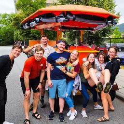 Eight friends smiling and laughing beside a bright orange pedal-powered party bike with a canopy on a sunny city street, trees and parked cars in the background