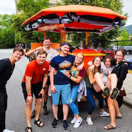Eight friends smiling and laughing beside a bright orange pedal-powered party bike with a canopy on a sunny city street, trees and parked cars in the background
