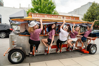 Six women laughing and pedaling a bright orange pedal pub on a downtown street, seated on bar-style saddles under a canopy during a daytime city tour.