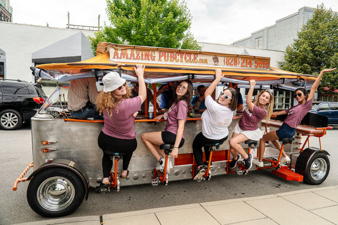Six women laughing and pedaling a bright orange pedal pub on a downtown street, seated on bar-style saddles under a canopy during a daytime city tour.