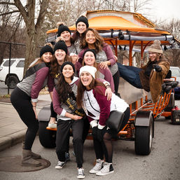 Smiling group of women in matching maroon-and-gray shirts and knit hats posing on an orange party bike on a winter urban street, bachelorette celebration vibe.