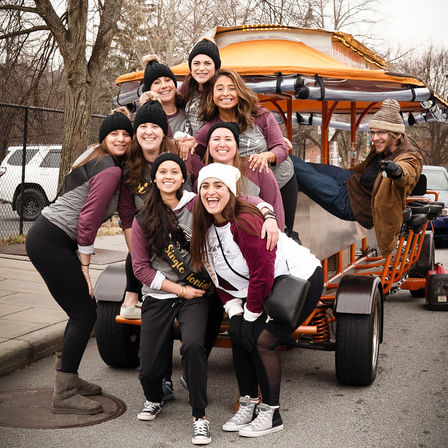 Smiling group of women in matching maroon-and-gray shirts and knit hats posing on an orange party bike on a winter urban street, bachelorette celebration vibe.
