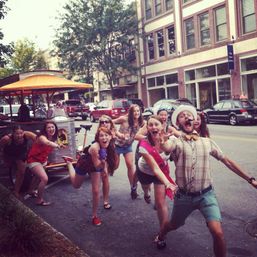 Cheerful group of friends running and laughing toward the camera on a downtown street beside a pedal pub, one wearing a pink bachelorette sash and another in a straw hat, with brick storefronts and parked cars in the background.