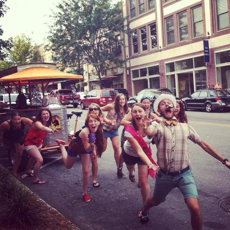Cheerful group of friends running and laughing toward the camera on a downtown street beside a pedal pub, one wearing a pink bachelorette sash and another in a straw hat, with brick storefronts and parked cars in the background.