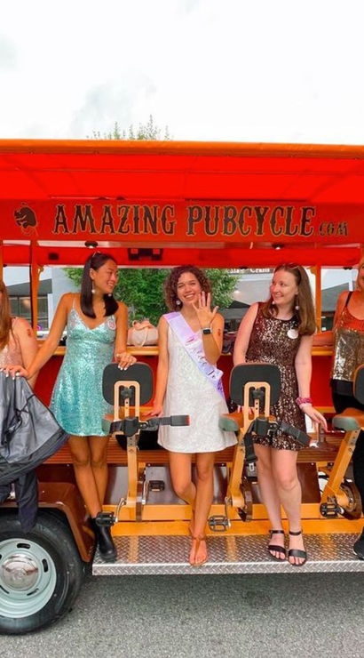 Six women in colorful, sparkly dresses laugh and pose aboard a bright orange pedal-powered party bike parked on a downtown street