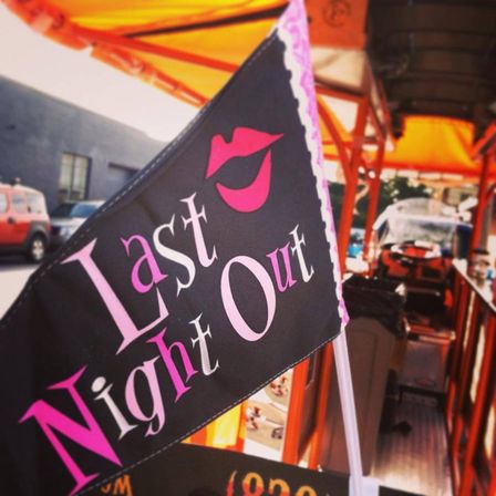 Black triangular flag with pink lips and the words "Last Night Out" attached to an orange pedal party bike on a sunny city street