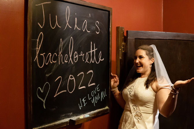 Smiling bride-to-be wearing a veil and sash poses next to a chalkboard sign reading bachelorette 2022 in a dimly lit indoor party venue.